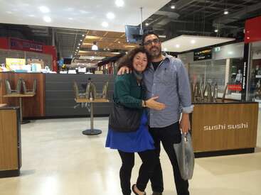 Das Bild zeigt einen Mann und eine Frau, die für ein Foto in einem Food Court posieren, mit einem Sushi-Stand und leeren Stühlen im Hintergrund.