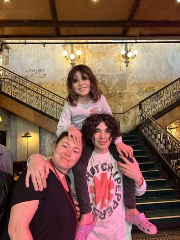 Das Bild zeigt eine Frau und einen Mann, die mit einem Kind auf den Schultern vor einer Treppe posieren, die vor einer Wand mit einem dekorativen Geländer steht.