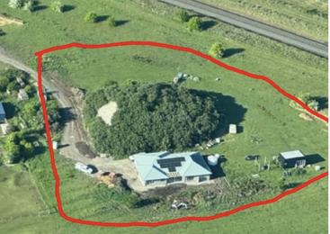 Dieses Luftbild zeigt ein Haus mit einem Solarmodul-Dach, das von Bäumen und offenem Grasland umgeben ist und von einer roten Begrenzungslinie umschlossen wird. In der Nähe sind Schuppen zu sehen.