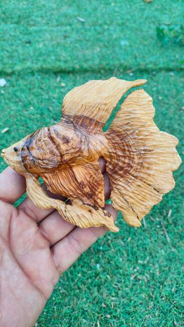 Eine Hand hält eine detaillierte Holzschnitzerei, die einem Betta-Fisch ähnelt, mit komplizierten Flossen und natürlicher Holzmaserung, vor einem verschwommenen grünen Grashintergrund.