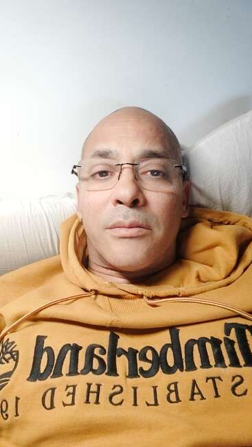 Eine Person mit Brille und einem gelben Timberland-Kapuzenpulli liegt auf einem weißen Kissen und schaut mit ruhigem, neutralem Blick direkt in die Kamera.