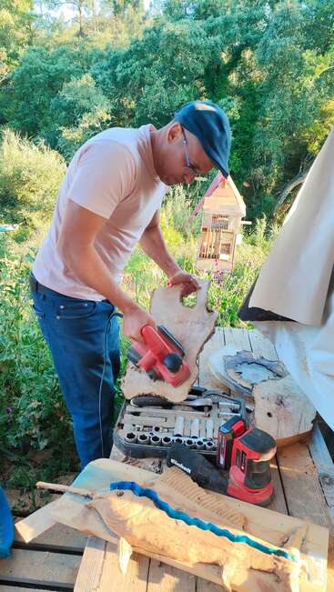 Ein Mann mit Brille und Mütze schleift im Freien, umgeben von Grün, ein Holzstück. In der Nähe befinden sich verschiedene Holzbearbeitungswerkzeuge und ein teilweise fertiges Projekt.