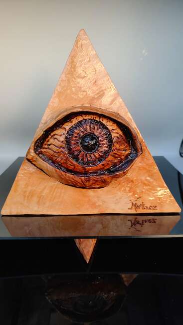 Dieses Bild zeigt eine dreieckige Skulptur mit einem realistischen, geschnitzten Auge in der Mitte. Auf dem Sockel sind die Namen "Martinez" und "Naypres" eingraviert.