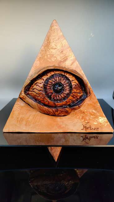 This image shows a triangular sculpture resembling an eye embedded in a pyramid. It has a realistic, wood-textured finish with artist signatures “Martinez” and “Vaquess.”