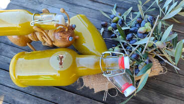 Drei Flaschen Olivenöl, eine geschnitzte Holzbiene und frische Olivenzweige mit Oliven werden auf einer rustikalen Holzfläche ausgestellt und sorgen für eine natürliche Atmosphäre.