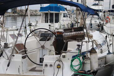 This image shows the cockpit of a sailboat docked at a marina. There are ropes, fenders, a large steering wheel, and nearby boats in the background.