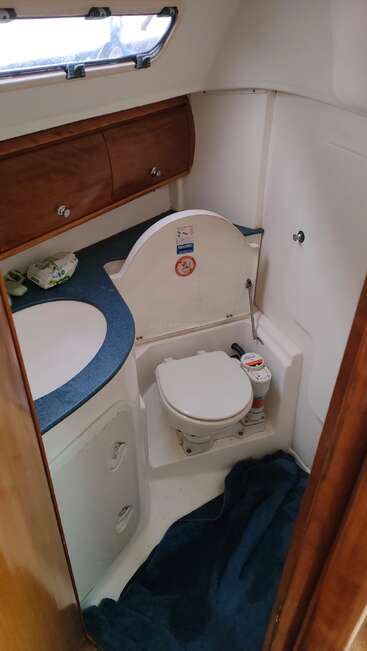 The image shows a compact boat bathroom with a marine toilet, blue countertop, sink, wooden storage cabinets, toiletries, a towel on the floor, and a small window.
