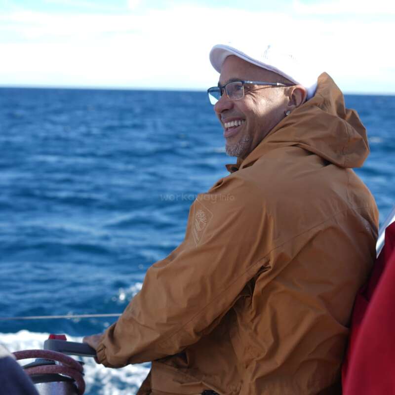 A smiling man wearing glasses, a brown jacket, and a white hat enjoys a boat ride on the ocean, with blue water and sky in the background.