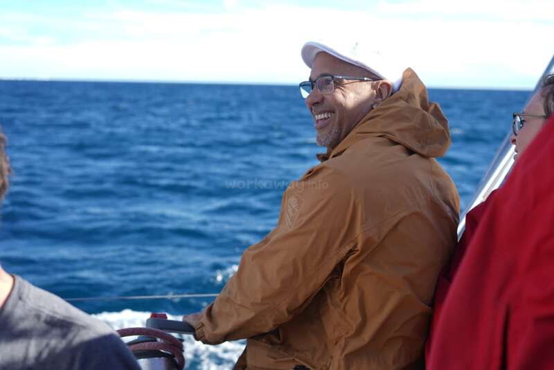 Ein lächelnder Mann mit Brille, brauner Jacke und weißem Hut genießt eine Bootsfahrt auf dem Meer, mit blauem Wasser und Himmel im Hintergrund.