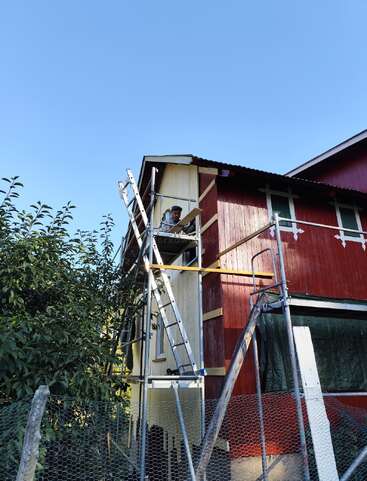 Ein Mann arbeitet auf einem Gerüst und streicht oder repariert den oberen Teil eines roten Holzhauses. Bei Tageslicht sind das Haus von blauem Himmel und Bäumen umgeben.