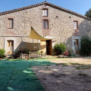 This charming stone house features large wooden doors and windows, a triangular sunshade, outdoor benches, green flooring, potted plants, and a relaxed rural atmosphere.