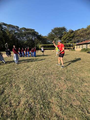 A group of students in uniforms watch an instructor holding a volleyball, preparing to serve on an outdoor grassy court, under a clear blue sky.
