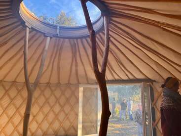 A luz do sol passa por uma claraboia circular dentro de uma iurta. Vigas de madeira sustentam a estrutura. Várias pessoas ficam do lado de fora, enquanto uma pessoa fica do lado de dentro da porta.