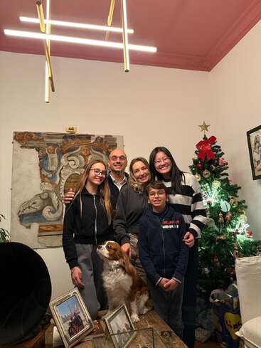 A happy family with three children and a dog pose together, smiling by a decorated Christmas tree. Framed photos and artwork decorate the cozy living room.