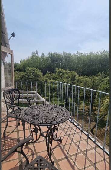 A sunlit balcony with ornate metal table and chairs overlooks lush green trees. Terracotta tiles and white railing create a peaceful, inviting outdoor seating area surrounded by nature.