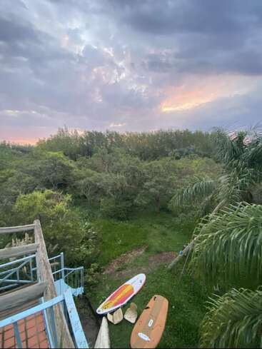The image shows a lush green landscape at sunset, with dramatic clouds, trees, palm leaves, surfboards, and a pastel-colored stair railing in the foreground. Peaceful scene.