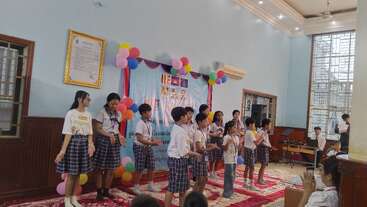 Un groupe d'enfants en uniforme se tient sur une scène décorée de ballons ; ils se produisent ou font une présentation devant un public dans la salle polyvalente d'une école.