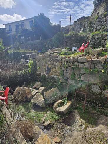 Un jardin en terrasse ensoleillé avec de vieux murs en pierre, des chaises rouges, des rochers éparpillés, une échelle rouillée, des plantes envahissantes et une maison en arrière-plan sous un ciel bleu.