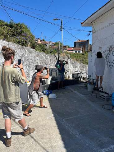 Trois hommes photographient une femme posant sur une chaise devant un mur blanc extérieur orné de graffitis. Il semble s'agir d'une séance photo informelle par une journée ensoleillée.