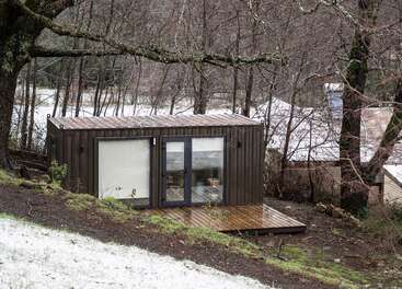 A modern tiny house made from a shipping container sits on a wooden deck amidst leafless trees, surrounded by light snow and natural, rustic scenery.