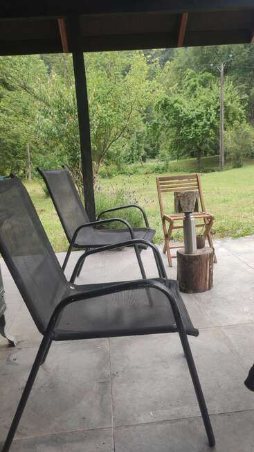 Two black mesh chairs and a wooden chair sit on a patio. A thermos and mate cup rest on a tree stump, surrounded by green trees and nature.