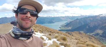 Ein Mann, der eine Sonnenbrille, einen Hut und eine Halskrause trägt, lächelt in die Kamera. Er steht im Freien, umgeben von verschneiten Bergen, Gras und einem See.