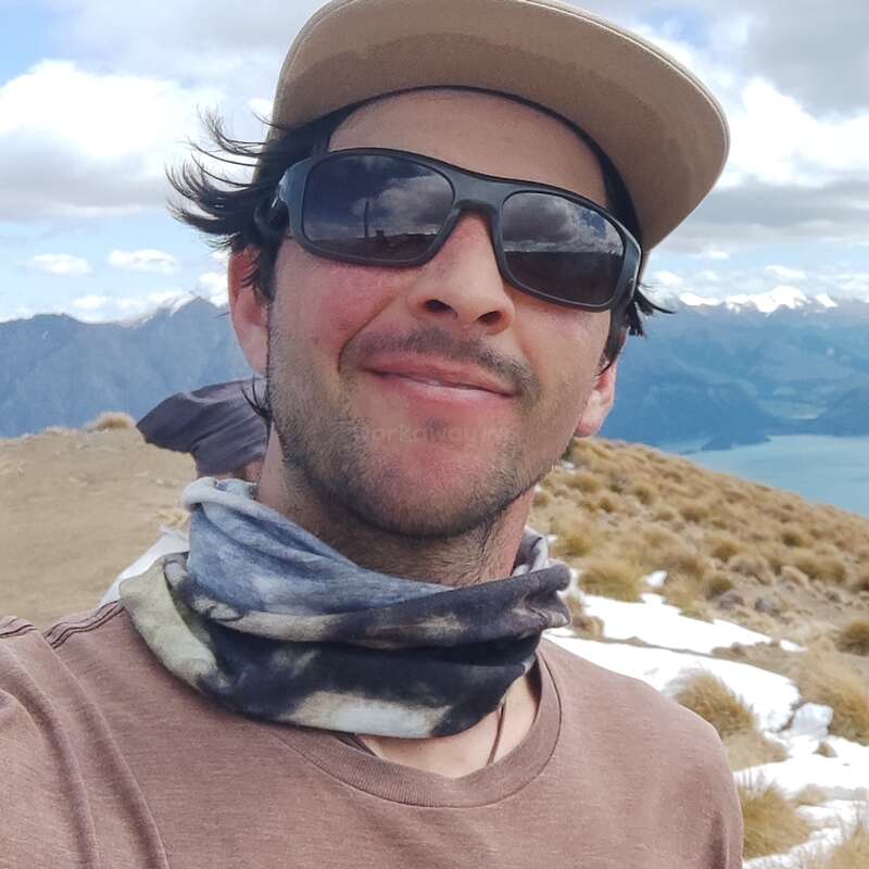 A man wearing sunglasses, a hat, and a neck gaiter smiles at the camera. He is outdoors, surrounded by snowy mountains, grass, and a lake.