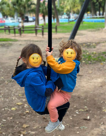 Zwei Kinder in blauen Jacken spielen zusammen auf einer Schaukel in einem Park. Ihre Gesichter sind zum Schutz der Privatsphäre mit Smiley-Emojis bedeckt.