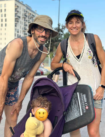 Ein fröhliches Paar posiert im Freien mit einem kleinen Kind in einem lila Kinderwagen. Das Gesicht des Kindes ist von einem Smiley-Emoji verdeckt, das ein Stofftier hält.