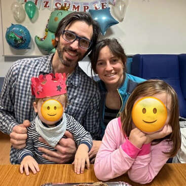 Eine vierköpfige Familie posiert für eine Geburtstagsfeier. Zwei Kinder haben Emoji-Gesichter, eines trägt eine Krone. Luftballons und Dekorationen bilden einen festlichen Hintergrund.