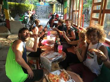 The image depicts a group of eight people enjoying pizza at a table in a restaurant, with a motorcycle visible outside through the windows.