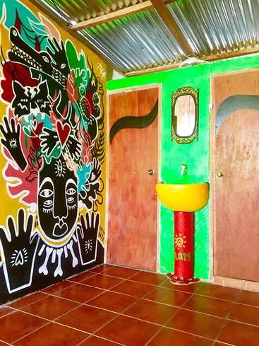 The image depicts a vibrant room with a mural on the left wall, a sink in the corner, and two doors, featuring a mix of bright colors and eclectic artwork.