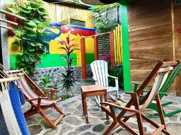 The image depicts a serene outdoor seating area with wooden chairs, a hammock, and a vibrant mural, exuding a tropical atmosphere.