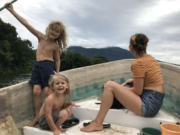 The image depicts three children on a boat, with one standing and two sitting, all with blonde hair, set against a scenic backdrop of trees and mountains.