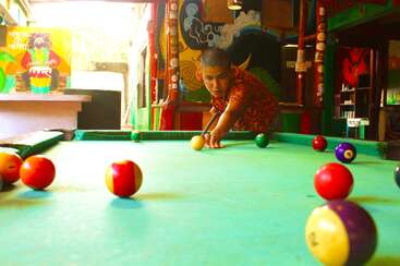 A person focuses intently while playing pool in a vibrant, colorful room adorned with murals and art. Bright billiard balls scatter across the green table.