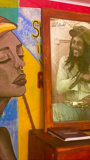 The image displays a colorful mural of a woman with closed eyes beside a framed photograph of a smiling man, both exuding vibrant artistic and cultural energy.