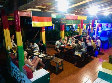 A group of people enjoy dinner and conversation in a colorful, lively outdoor restaurant with painted pillars, German flags, hammocks, and vibrant lighting at night.