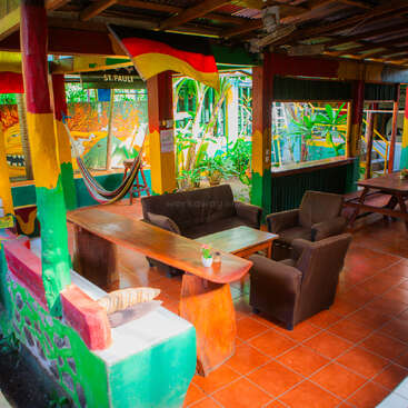 Esse vibrante e colorido lounge ao ar livre conta com assentos aconchegantes, redes, murais animados, mesas de madeira e plantas tropicais, criando um ambiente descontraído, artístico e acolhedor para os hóspedes.
