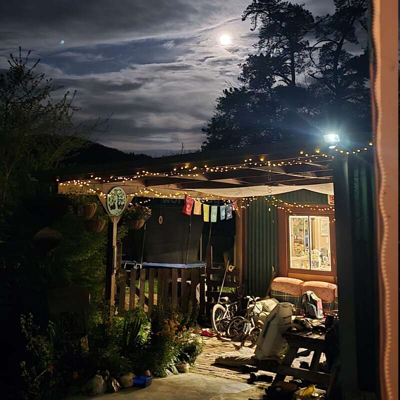 Eine gemütliche, beleuchtete Veranda mit Lichterketten hängt unter einem wolkigen Nachthimmel. Der Vollmond leuchtet über den Silhouetten der Bäume; Fahrräder und Gartenpflanzen liegen ruhig darunter.