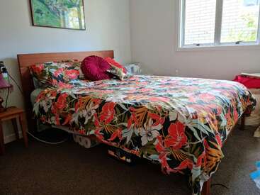 La imagen muestra un dormitorio con una cama con un vibrante edredón floral, un cabecero de madera y una ventana con un marco blanco, sobre una pared blanca.