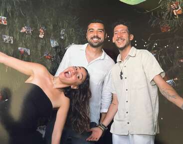 La imagen muestra a tres individuos posando para una foto, con dos hombres de pie detrás de una mujer que está inclinada hacia atrás y extiende el brazo hacia un lado.