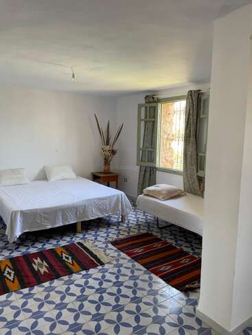 Esta habitación cuenta con una cama doble y una cama individual, alfombras con estampados de colores, suelo de baldosas geométricas, una ventana abierta, una mesilla de noche rústica y plantas secas decorando la esquina.