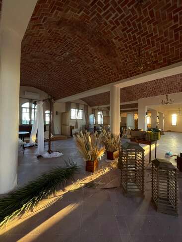 Una sala espaciosa con techos de ladrillo arqueados, pilares blancos, grandes ventanales, luz natural, plantas decorativas, farolillos y algunos materiales dispersos, que dan un ambiente rústico y cálido.