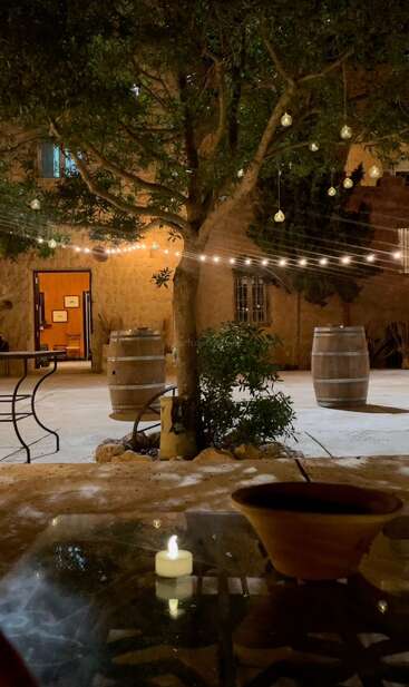 Un acogedor patio exterior por la noche, decorado con luces de cuerda y bombillas colgantes, barriles rústicos, un árbol y una mesa con un cuenco a la luz de las velas. Ambiente tranquilo.