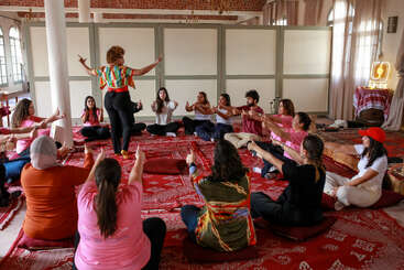 A group of people sit in a circle on red cushions, following an instructor's movements in a spacious, warmly lit room with patterned rugs and soft lighting.