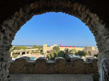 Framed by a stone archway, the image reveals a scenic view of a Mediterranean castle courtyard, lush greenery, bright blue sky, and tranquil sea beyond.