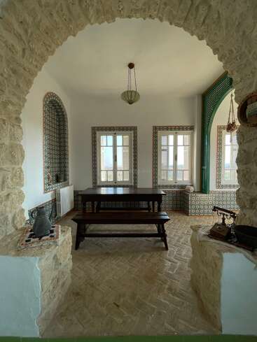 This cozy room features rustic stone arches, patterned tiles, a wooden dining table with benches, vintage décor, twin windows, and a hanging lamp, evoking Mediterranean charm.