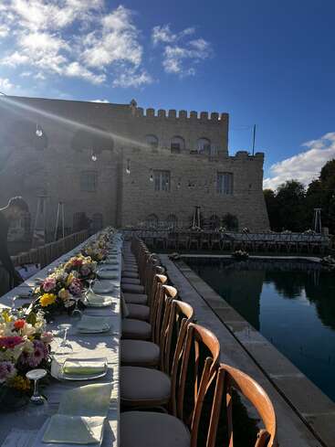 A long, elegant dining table set with flowers by a reflective pool. Castle-like stone building in the background, under a bright blue sky.