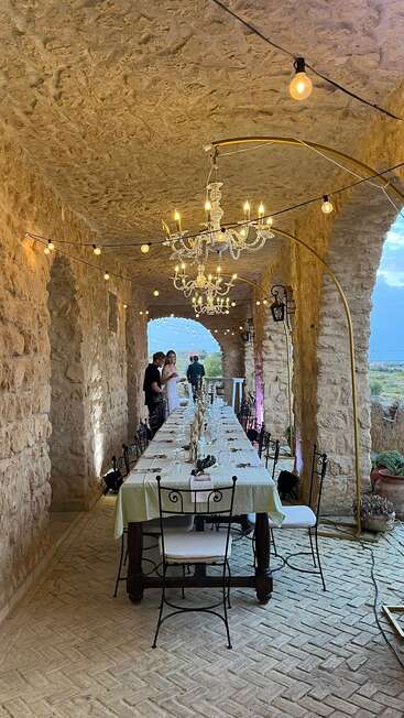A long dining table set for an event under a rustic stone archway, with chandeliers and string lights, guests chatting, and scenic countryside view outside.