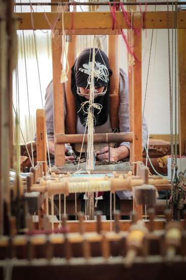 A person wearing a headscarf is focused on weaving fabric using a traditional wooden loom, surrounded by threads, demonstrating a handcrafted textile-making process.